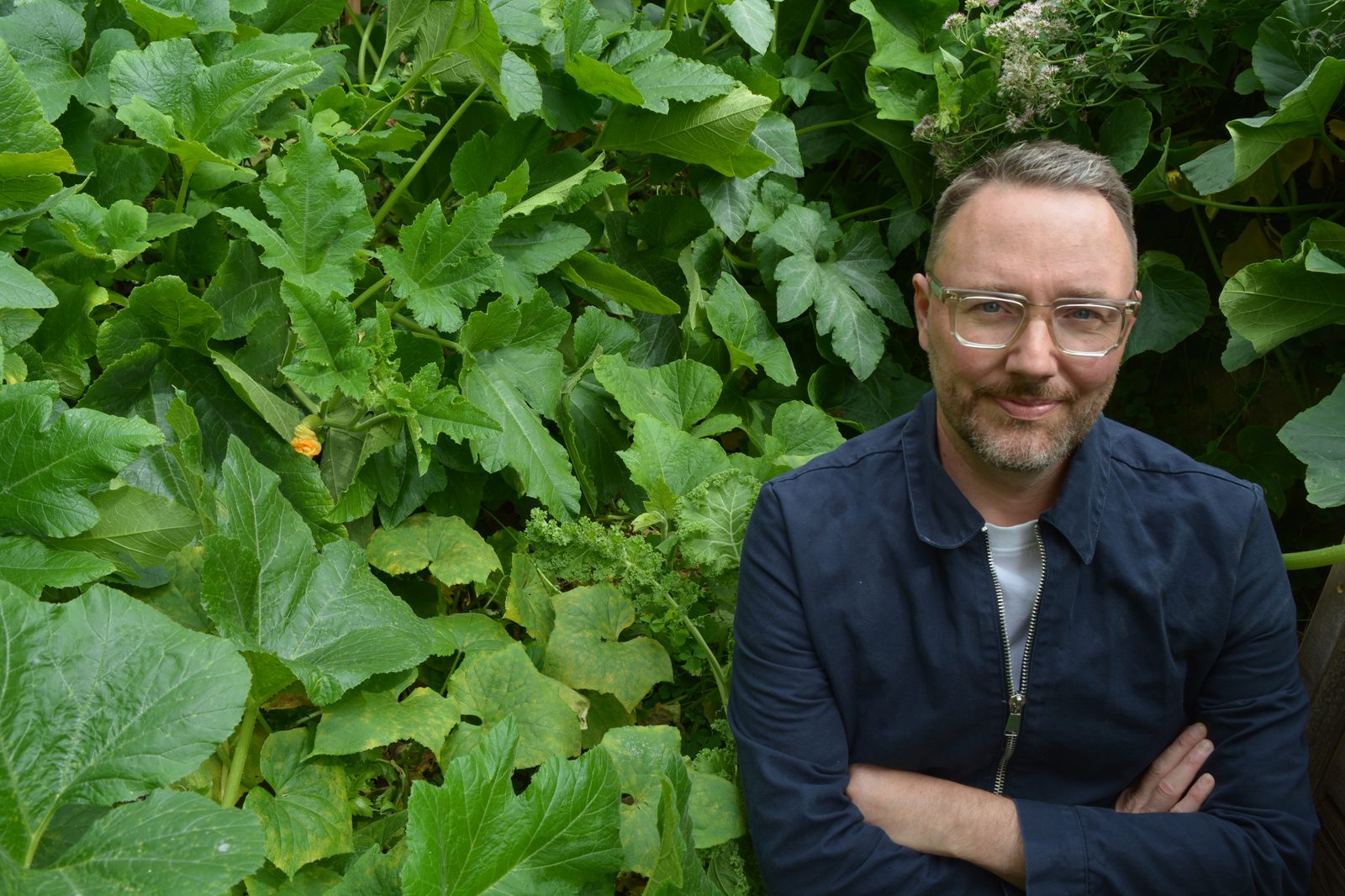David Ruffieux, bras croisés, lunettes claires et sourire discret, adossé à une végétation potagère luxuriante